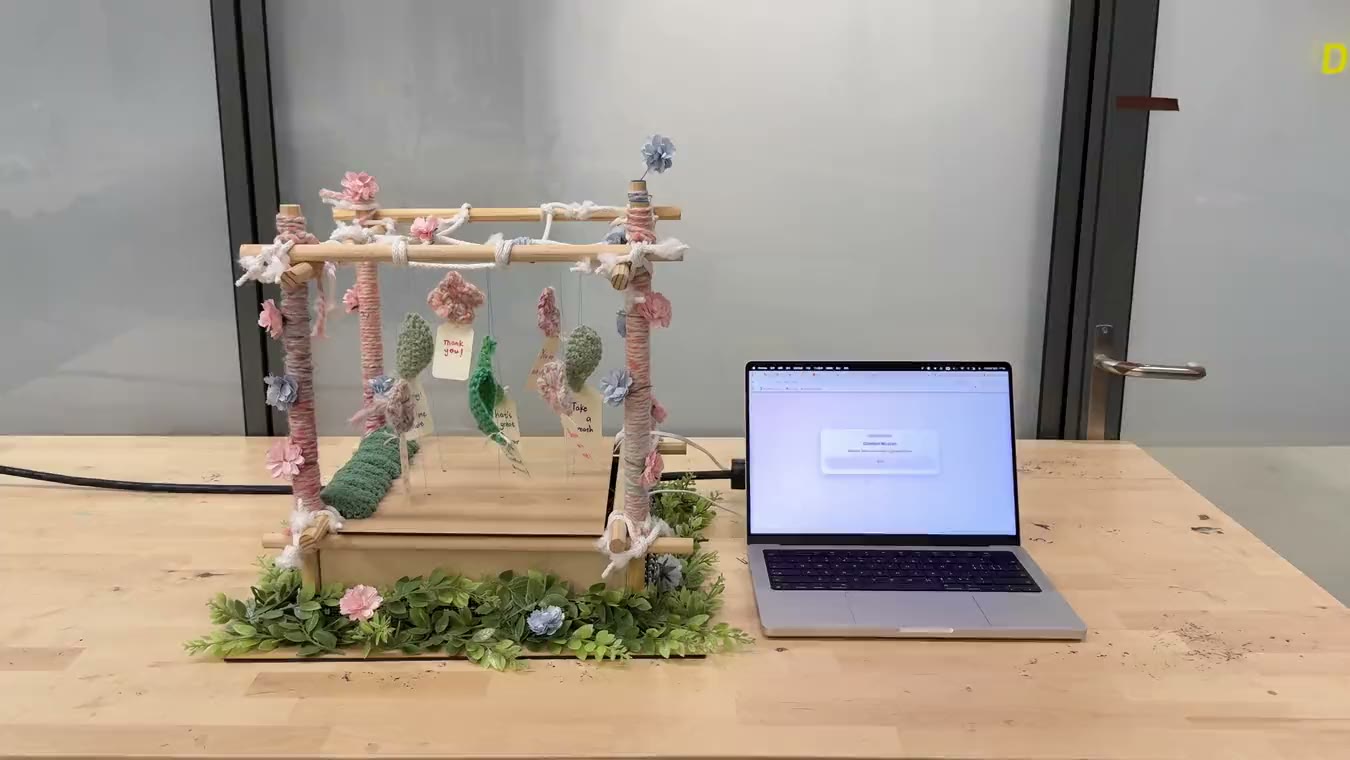 A hanging mobile of small fabric flowers on a wooden frame, next to a laptop on a wooden table